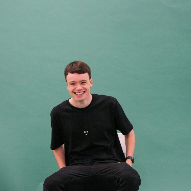 Smiling young man in a black shirt seated against a green background.