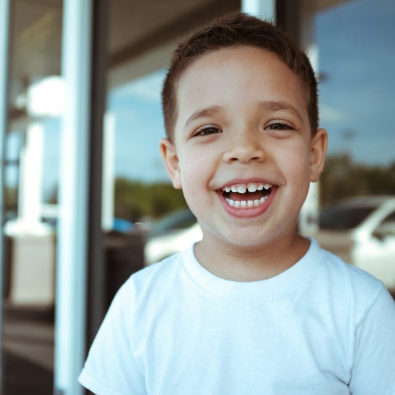 Smiling young boy wearing a white shirt, standing outside with cars in the background.